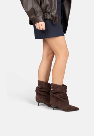 Woman wearing a dark brown leather jacket, navy blue skirt, and slouchy dark brown suede ankle boots with low heels, standing sideways.