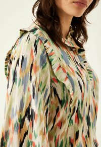 Blusa colorata e a fantasia con colletto arricciato, realizzata in tessuto leggero con tonalità di verde, blu, arancione e accenti neri.
