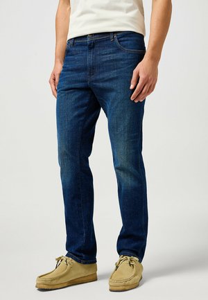 Blue denim jeans with a straight fit, five-pocket design, and subtle fading. Worn with tan moccasin-style shoes on a light background.