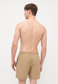 Shorts de bain beiges avec une taille élastique, des poches latérales et une texture lisse, vus de dos avec un torse nu visible.