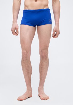 Mužský model stojaci naboso v modrých priliehavých plavkách na jednoduchom svetlosivom pozadí.