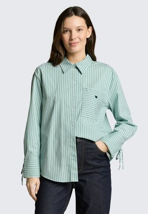 Młoda kobieta w jasnozielonej koszuli w paski z kieszenią na piersi i ciemnoniebieskich dżinsach, stojąca z jedną ręką w kieszeni.
