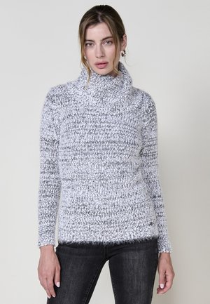 Cowl-Neck-Pullover in Hellgrau mit strukturiertem Strickmuster. Weicher Stoff mit leicht flauschiger Oberfläche. Kombiniert mit dunklen Jeans.