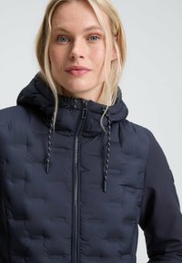 Donna bionda con occhi azzurri che indossa una giacca trapuntata blu navy con cappuccio e lacci su uno sfondo chiaro e uniforme.