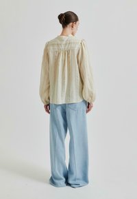 Svetlo bež bluzica z navpičnimi črtami in nabranimi rokavi, v kombinaciji z visokimi širokimi svetlo modrimi kavbojkami. Enostavna ozadje.