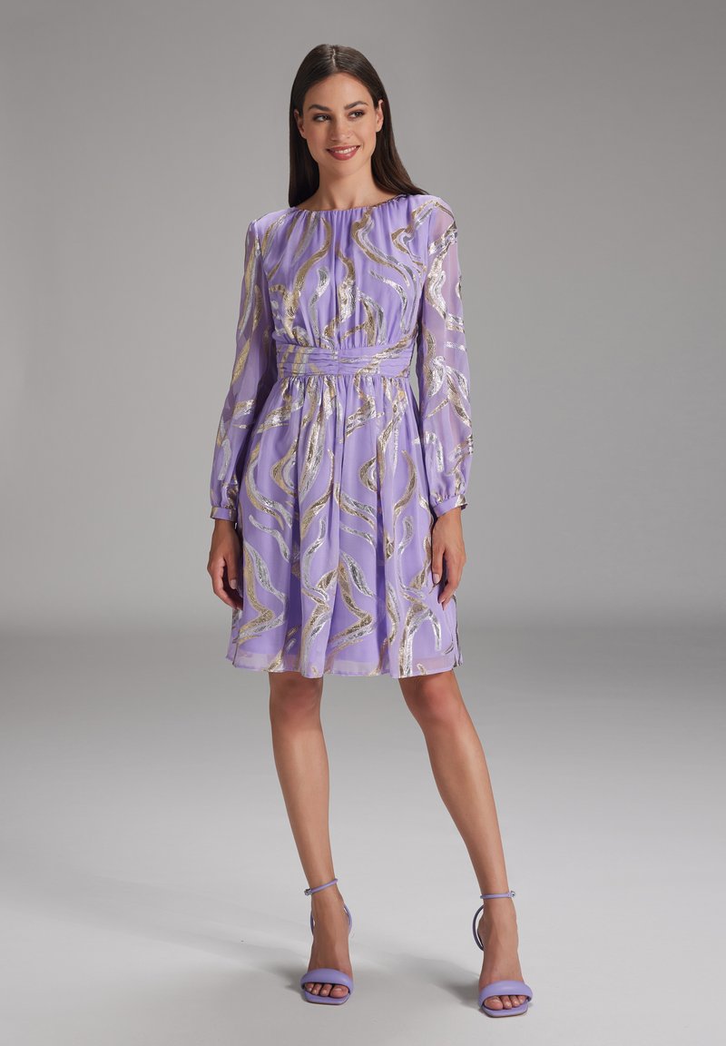 Robe lavande avec des manches longues, motif tourbillonnant doré, taille cintrée et jupe fluide. Assortie à des talons lavande.