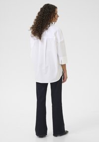 Camicia bianca oversize con maniche a tre quarti, orlo stondato e spacchi laterali; abbinata a jeans svasati scuri. Tessuto morbido al tatto.