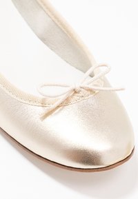 Bailarina metálica dorada con punta redondeada, ribete de tela en el tobillo y un lazo anudado en la parte frontal como detalle. Textura suave en general.