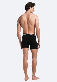 Svarta bomullsnäbbekalsonger med ett vitt varumärkes-bälte, visade bakifrån på en modell med bara fötter. Enkelt, figurnära design.