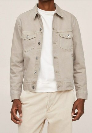 Mężczyzna w beżowej, zapinanej na guziki kurtce jeansowej, białej bluzie i pasujących beżowych spodniach, stoi z rękami swobodnie opuszczonymi wzdłuż ciała.