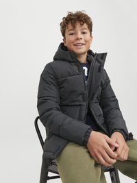 Black puffer jacket with a hood, featuring a zip closure and elastic cuffs, paired with green trousers, on a chair against a plain background.