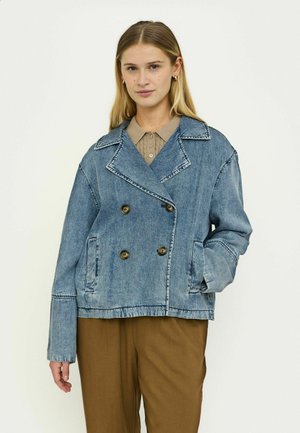 Jeansjacke in Hellblau, zweireihiges Design mit großen Knöpfen, Reverskragen und Seitentaschen. Trägt sich über einem beigen Hemd mit Kragen.