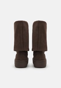 Pari korkeita, tummanruskeita mokkasiinibootseja, joissa on litteät, paksut pohjat, esitelty takaa valkoista taustaa vasten.