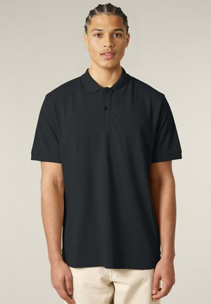 Jonge man met gevlochten haar, gekleed in een eenvoudige zwarte polo met korte mouwen en lichtgekleurde broek, staande voor een neutrale achtergrond.