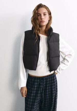 Jeune femme aux cheveux ondulés portant un gilet matelassé noir par-dessus une chemise blanche à manches longues et un pantalon à carreaux foncé, debout sur un fond blanc.
