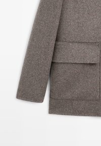 Cappotto in lana grigia con finitura leggermente strutturata, caratterizzato da una grande tasca frontale, maniche ampie e un design a taglio dritto.