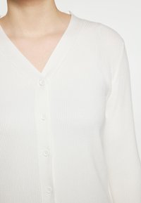 Weißer gerippter Cardigan mit V-Ausschnitt, der sechs passende Knöpfe auf der Vorderseite hat. Der Stoff wirkt weich und leicht.