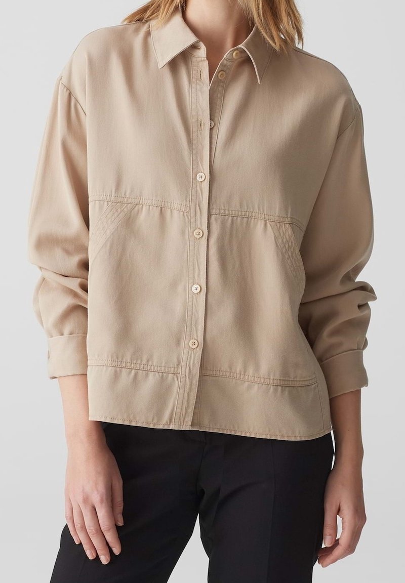 Femme portant une chemise beige à manches longues boutonnée avec un pantalon noir, manches retroussées, debout devant un fond uni.