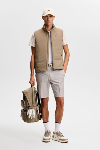 Άνδρας με μπεζ καπέλο, αμάνικο γιλέκο, λευκό μπλουζάκι, σορτς με σχέδια, κρατώντας μπεζ σακίδιο με λευκά γάντια, στέκεται μπροστά σε απλό φόντο.