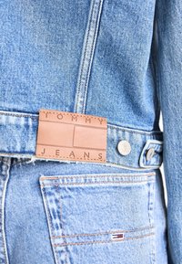 Chaqueta de mezclilla azul claro con un parche de cuero marrón grabado con "TOMMY JEANS" y detalles de botones de metal. Bordes deshilachados visibles.