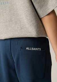 Tumši zili sporta bikses ar sānu kabatu, kurām ir balts "ALLSAINTS" logo. Mīksta kokvilnas auduma ar gludu tekstūru un pielāgotu piegriezumu.