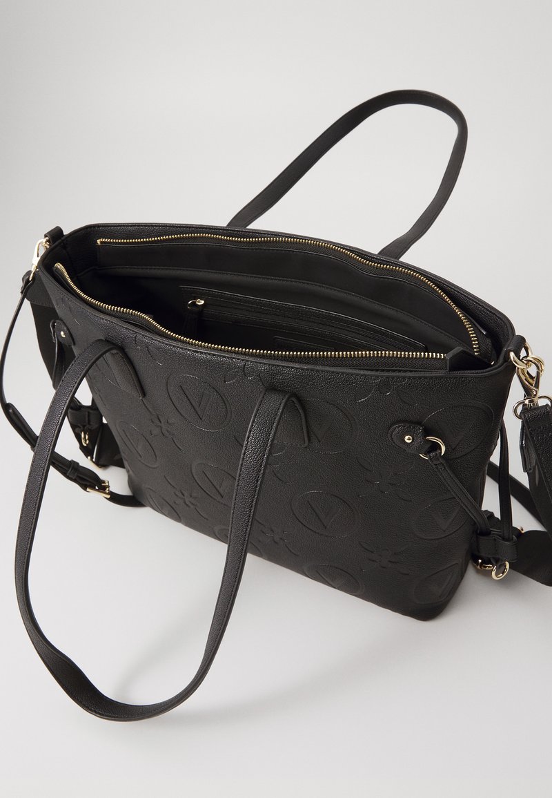 Borsa tote sintetica nera con motivo logo in rilievo, doppi manici superiori, chiusura con zip e scomparti interni. Accenti in hardware color oro.