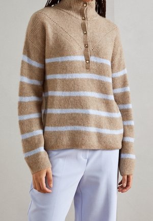 Pull beige en tricot avec des rayures horizontales blanches, col montant et boutons sur le devant, porté avec un pantalon clair.