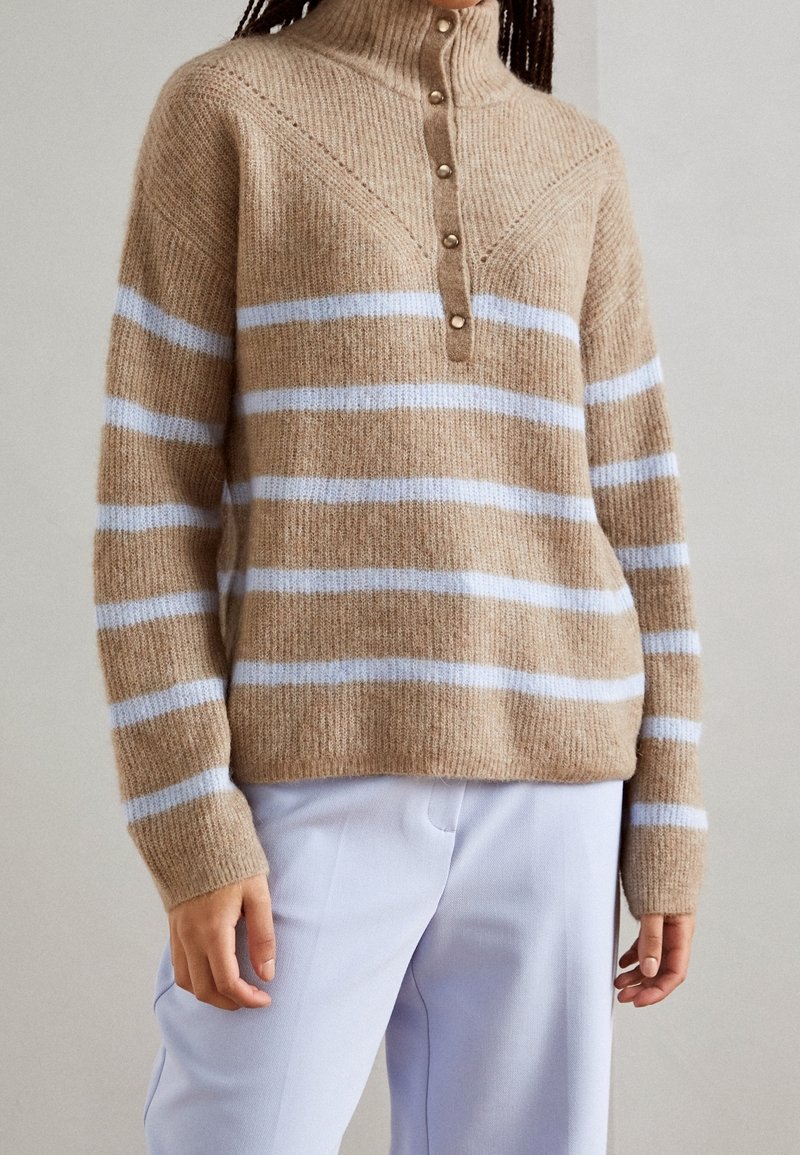 Pull beige en tricot avec des rayures horizontales blanches, col montant et boutons sur le devant, porté avec un pantalon clair.