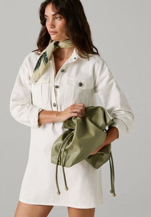 Žena u bijeloj haljini s gumbima i šal s uzorkom oko vrata drži velikanu torbu maslinasto zelene boje uz sivo pozadinu.