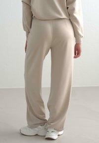 Beige Sweatpants mit lockerer Passform, die eine glatte Textur aufweisen, einen elastischen Bund haben und mit weißen Sneakern auf einem neutralen Boden kombiniert sind.