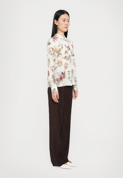 Bluse mit floralem Muster und langen Ärmeln in Creme, kombiniert mit dunkelbraunen geplissten Hosen. Das Model trägt weiße Slipper.