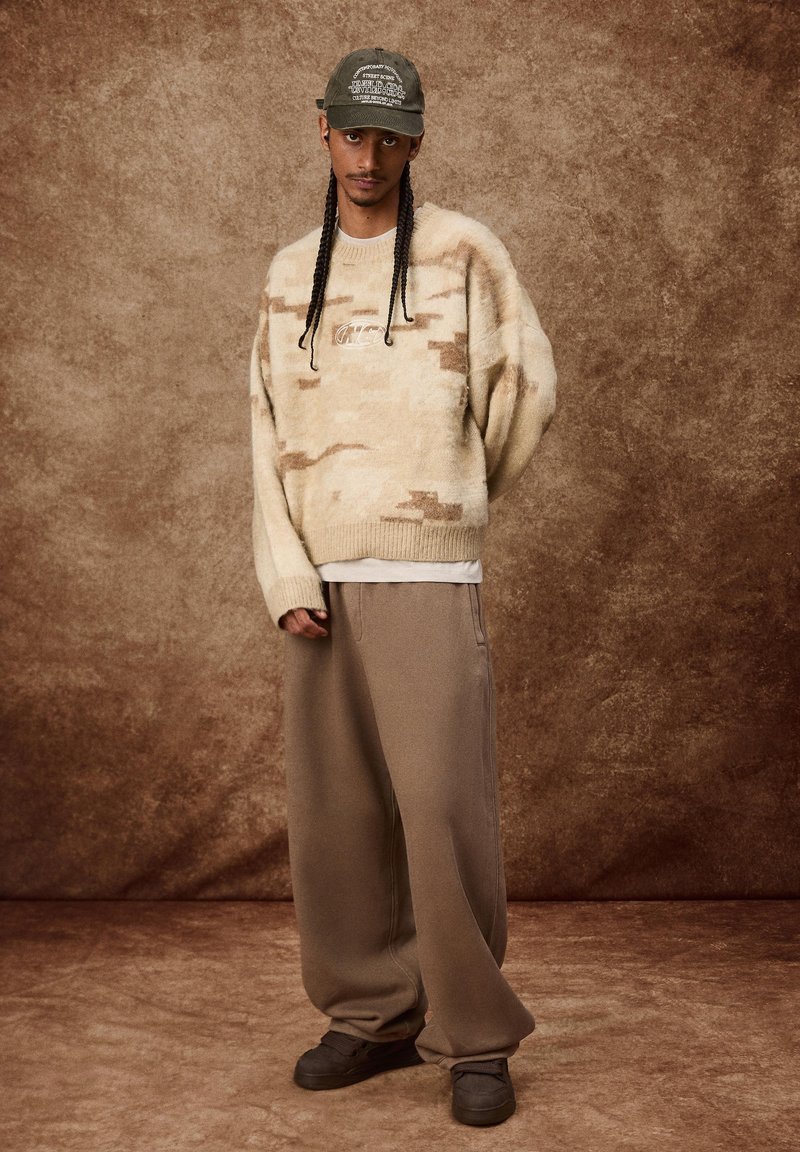 Beige, kamouflagemönstrad tjock tröja, ljus t-shirt under, lösa bruna joggingbyxor och mörka sneakers. Bärs med en grön keps.