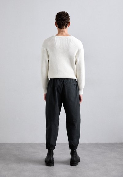 Valkoinen ribattu neule, mustat joustovyötäröiset housut, joissa on lahkeensuukappaleet, yhdistetty mustiin nilkkureihin. Neutraali tausta täydentää asua.