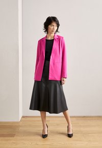 Bright pink blazer over a black top, paired with a black leather midi skirt and black high heels, standing on wooden floor.