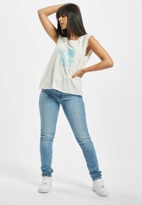 Lys grå ermeløs topp med blå grafisk tekst, kombinert med tettsittende lyseblå jeans og hvite joggesko, alt på en enkel hvit bakgrunn.