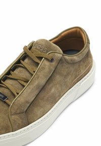 Sneaker in suede marrone con punta arrotondata, lacci oliva e suola in gomma bianca. Presenta un logo impresso e dettagli di cucitura sottili.