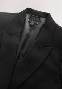 Black blazer with a satin finish, sharp lapels, and a single front pocket. Features a branded interior label. Smooth texture.
