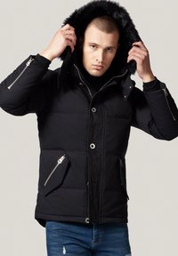 Czarna kurtka puchowa z odpinanym kapturem wyłożonym futrem, z metalowymi zamkami błyskawicznymi, zatrzaskami oraz kieszeniami bocznymi. Miękka tekstura i pikowany design.