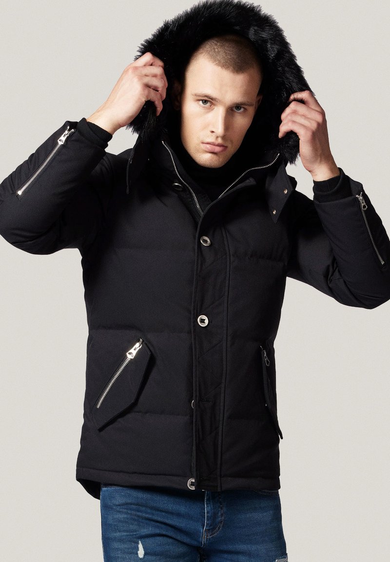 Czarna kurtka puchowa z odpinanym kapturem wyłożonym futrem, z metalowymi zamkami błyskawicznymi, zatrzaskami oraz kieszeniami bocznymi. Miękka tekstura i pikowany design.