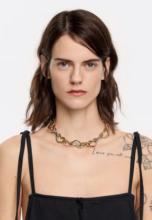 Gold, silver, and rose gold heart-shaped metal necklace with interlinked chains, worn on a bare shoulder over a black top.