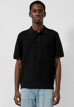 Man draagt een zwarte, korte mouw gebreide poloshirt en lichtblauwe spijkerbroek, staand tegen een effen lichte achtergrond.