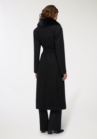 Cappotto di lana nera con colletto in pelliccia sintetica, maniche lunghe, vita con cintura e taglio dritto. Presenta bottoni ai polsi e una texture liscia.