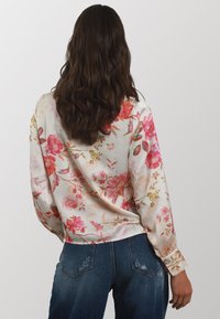 Blusa in satin floreale con maniche lunghe, caratterizzata da motivi floreali rosa e rossi su uno sfondo crema, abbinata a jeans strappati blu.