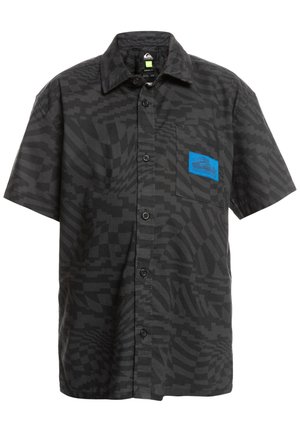 Enfant debout, les mains sur les hanches, portant une chemise noire à manches courtes à motifs avec boutons, un short bleu clair, des chaussettes blanches et des baskets beige avec lacets noirs.