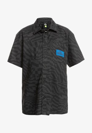 Enfant debout, les mains sur les hanches, portant une chemise noire à manches courtes à motifs avec boutons, un short bleu clair, des chaussettes blanches et des baskets beige avec lacets noirs.