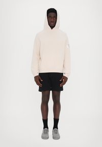 Light beige hoodie with a front pocket and logo patch, paired with black shorts and gray sneakers, on a plain white background.
