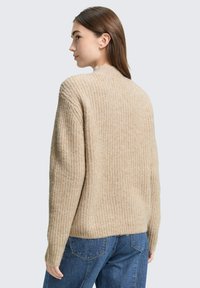 Beige gestrickter Pullover mit hohem Kragen, geripptem Muster und lockerer Passform, kombiniert mit blauen Jeans.