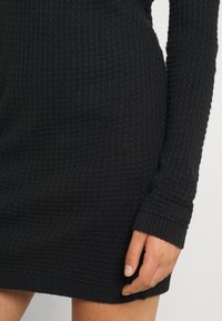 Robe en maille côtelée noire à manches longues, présentant un motif texturé et une silhouette ajustée. L'ourlet se situe au-dessus du genou.