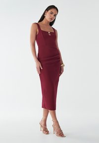 Burgundy tætsiddende kjole med firkantet udskæring, nøglehul detalje og samlede sider, parret med gyldne strophæle. Glat stof struktur.