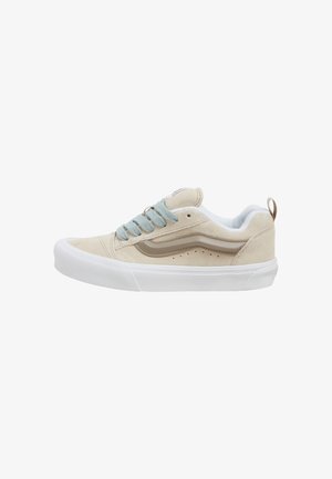 Beige Wildleder Low-Top Sneaker mit weißer Gummisohle, hellblauen Schnürsenkeln und beiger Seitenstreifen, auf einem weißen Hintergrund präsentiert.